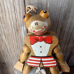 May include: Wooden frog puppet with a red bow tie, white shirt, and red and white striped shorts. The frog has large orange eyes and a smiling mouth. A string is attached to the top for hanging.