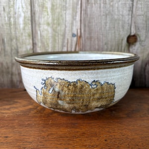 May include: A ceramic bowl with a cream-colored exterior and a brown rim. The bowl features a large, abstract brown design on the side. The bowl is sitting on a wooden surface.