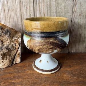 May include: A ceramic goblet featuring a wide, speckled mustard-yellow rim, a dark brown band, and a lower body with mottled brown and cream-colored glaze. The vessel stands on a pedestal base with a speckled white finish and a dark brown ring.