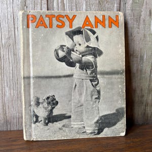 May include: Vintage book cover titled "PATSY ANN" in orange lettering. The black and white image shows a child in overalls and a sun hat, holding a ball, with a small dog nearby. The book has a worn appearance.