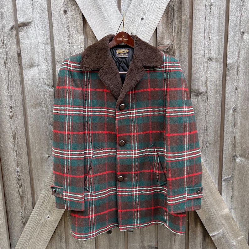 May include: A vintage plaid coat in shades of green, red, and brown, with a faux fur collar. The coat features a button-up front, two front pockets, and buttoned cuffs. The coat is hanging on a wooden hanger against a wooden background.