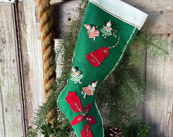 May include: A green felt Christmas stocking featuring a white cuff, decorated with small appliqued figures in striped clothing, a red bell shape, and red poinsettia leaves with gold sequins. The stocking is trimmed with gold piping.