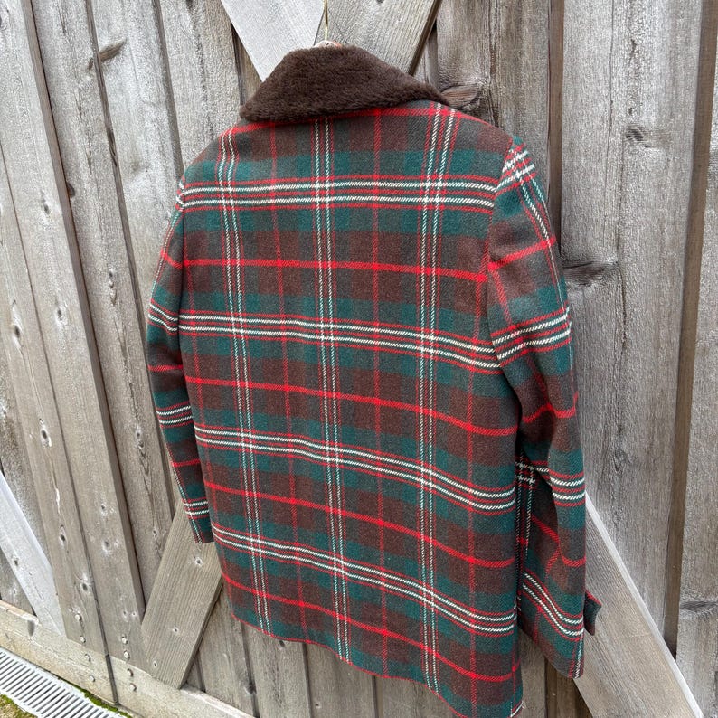 May include: A vintage plaid jacket in shades of green, red, brown, and white. The jacket features a classic plaid pattern and a brown faux fur collar. The jacket is hanging on a wooden surface.
