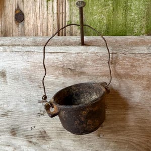 May include: A small, dark, weathered cast iron pot with a rounded body and two side handles. A thin, bent metal wire handle is attached to the sides, allowing the pot to hang from a single large, flat-headed metal nail driven into the wood.