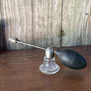 May include: A vintage atomizer featuring a clear glass base, a metallic silver-toned nozzle assembly with a pointed tip, and a black rubber bulb. The bulb has embossed text that reads DeVilbiss, and the device is designed for dispensing liquids.
