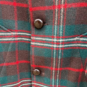 May include: Close-up of a plaid fabric with a pattern of red, green, white, and brown. The fabric has a textured appearance and is buttoned with two brown buttons. The garment appears to be a vest or jacket.
