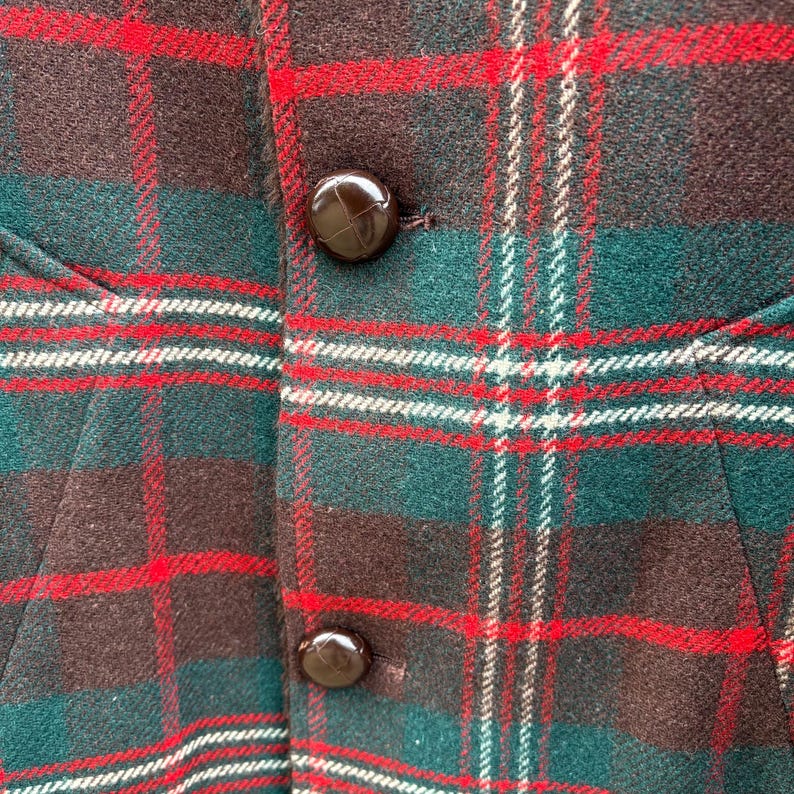 May include: Close-up of a plaid fabric with a pattern of red, green, white, and brown. The fabric has a textured appearance and is buttoned with two brown buttons. The garment appears to be a vest or jacket.