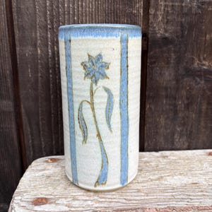 May include: A cylindrical ceramic vase with a light beige base, adorned with a blue floral design and vertical blue stripes. The rim is also blue. The vase is likely handmade and suitable for displaying flowers.