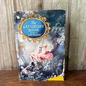 May include: Vintage hardcover book titled "The Saturday Book" edited by John Hadfield. The cover features a painting of a woman on a swing, with a blue and gold ornate frame around the title.