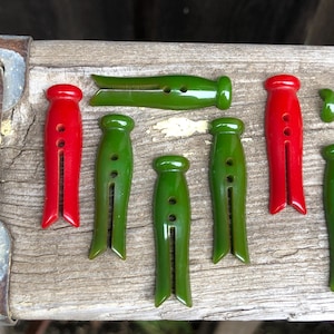 May include: A collection of vintage wooden clothespins in red and green. The clothespins are arranged on a weathered wooden surface. The clothespins have a classic design with a rounded top and a split bottom. The clothespins are approximately 3 inches long.