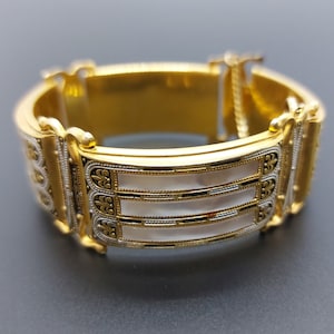 May include: A gold bangle bracelet with a white enamel inlay and intricate detailing. The bracelet has a hinged clasp.