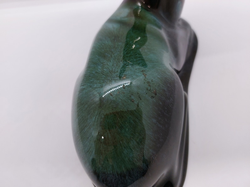 May include: A ceramic deer figurine with a dark green and black glaze. The deer is sitting down with its head tilted to the side. The glaze has a mottled effect, with darker green and black areas.