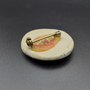 以下が含まれることがあります： ピンクとゴールドのデザインが施された白いセラミック製のブローチ。ブローチはピン留めです。