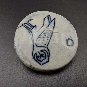 以下が含まれることがあります： 白いセラミック製の丸いピンで、青と白のイラストが描かれています。イラストは翼を広げたフクロウで、右を向いています。ピンの中央には、白い文字「O」が書かれた青い円があります。