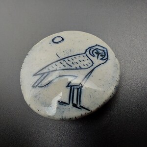 以下が含まれることがあります： 青と白のフクロウのイラストが描かれた白い丸いセラミックディスク。フクロウは右を向いており、詳細なデザインが施されています。