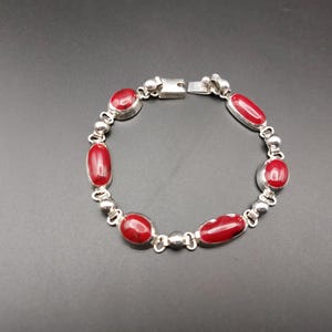 May include: Silver bracelet featuring oval and round red stones. The bracelet has a clasp closure and is composed of small silver beads and links. The jewelry is displayed on a dark surface.