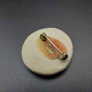 以下が含まれることがあります： 白い背景に桃色のデザインが施された丸いセラミック製のブローチ。ブローチの裏には金色のピンが付いています。