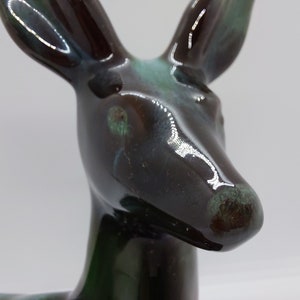 May include: A ceramic deer figurine with a glossy black and green glaze. The deer's head is tilted slightly to the side, and its eyes are closed. The glaze has a mottled appearance, with shades of brown, green, and blue.