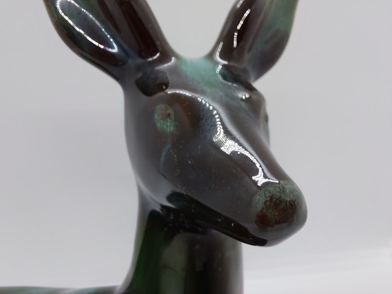 May include: A ceramic deer figurine with a glossy black and green glaze. The deer's head is tilted slightly to the side, and its eyes are closed. The glaze has a mottled appearance, with shades of brown, green, and blue.