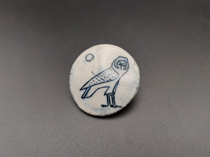 以下が含まれることがあります： 背景に円形の模様が入った、青い輪郭のフクロウが描かれた白いセラミック製のピン