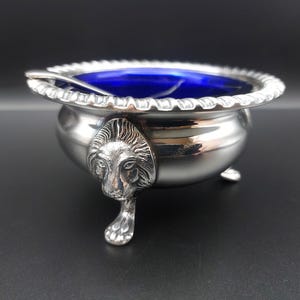 May include: A silver salt cellar with a cobalt blue interior and a small spoon. The bowl has a decorative rim and is supported by three small feet. A lion's head adorns the side of the bowl.