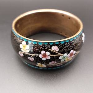 May include: A black bangle bracelet with a floral design. The bracelet is decorated with white, pink, and yellow flowers on a black background with a turquoise border.