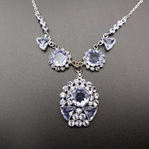 May include: A silver-toned necklace featuring a pendant and two floral-shaped accents, all adorned with light blue gemstones. The pendant is a cluster of stones, with triangular and round gems. The necklace chain is delicate and has matching gemstones.