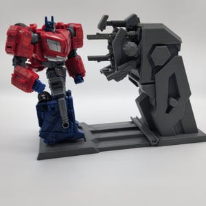 Puede incluir: Una figura de acci&oacute;n de robot rojo y azul se encuentra en una plataforma gris frente a un robot gris con m&uacute;ltiples armas. El robot con armas est&aacute; en una plataforma gris.