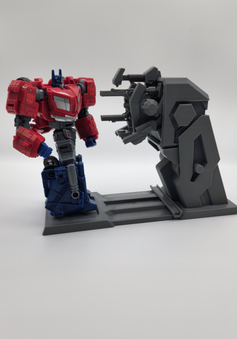 Puede incluir: Una figura de acci&oacute;n de robot rojo y azul se encuentra en una plataforma gris frente a un robot gris con m&uacute;ltiples armas. El robot con armas est&aacute; en una plataforma gris.