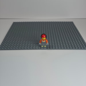 May include: A Lego construction worker minifigure with an orange vest, gray overalls, and a red hard hat stands on a large gray Lego baseplate. The baseplate is covered in small, raised studs.