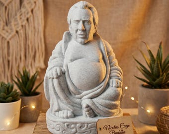 May include: A gray stone statue of Nicolas Cage in a seated Buddha pose. The statue has a detailed face and is on a wooden base. A small wooden tag reads "Nicolas Cage Buddha".