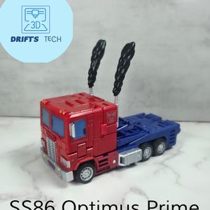 Może przedstawiać: Czerwono-niebieska zabawka ciężarówka Optimus Prime z czarnymi kominami dymnymi. Zabawka jest częścią zestawu ulepszeń Transformers o nazwie "SS86 Optimus Prime Smoke Upgrade Kit".