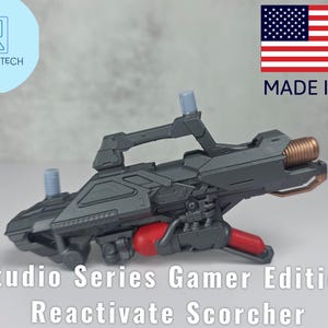 Può includere: Una pistola giocattolo Reactivate Scorcher Studio Series Gamer Edition grigio scuro con dettagli rossi e rame. La pistola giocattolo ha un'impugnatura, due attacchi cilindrici e la scritta "Made in USA". Il logo Drifts Tech è in alto a sinistra.