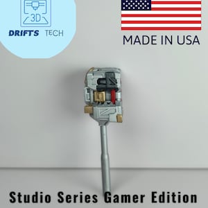 Puede incluir: Una maza en miniatura plateada y dorada con una cabeza detallada y un mango largo. La imagen incluye el texto "Studio Series Gamer Edition Reactivate Mini Gravity Mace" y un gráfico "Made in USA".