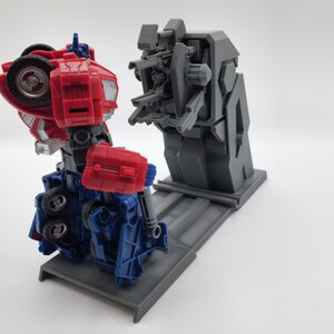 Puede incluir: Un juguete robot rojo y azul en modo robot est&aacute; de pie sobre una plataforma gris. El robot est&aacute; frente a una estructura gris con m&uacute;ltiples brazos y una base gris.