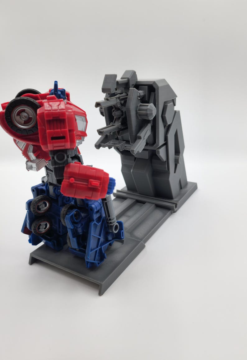 Puede incluir: Un juguete robot rojo y azul en modo robot est&aacute; de pie sobre una plataforma gris. El robot est&aacute; frente a una estructura gris con m&uacute;ltiples brazos y una base gris.