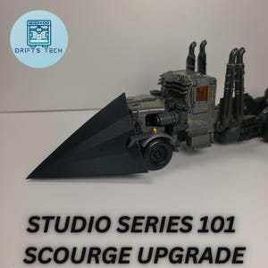 Op de afbeelding: Een grijze en zwarte speelgoedvrachtwagen met een groot zwart blad aan de voorkant. De tekst "STUDIO SERIES 101 SCOURGE UPGRADE" staat onderaan de afbeelding. Het logo "DRIFTS TECH" staat linksboven.
