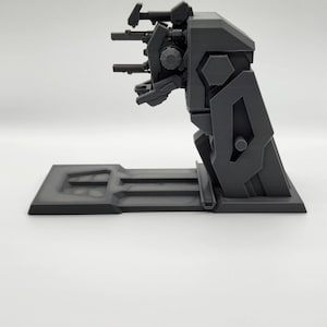 Puede incluir: Un modelo de pl&aacute;stico negro de un arma futurista con una torreta giratoria y varios ca&ntilde;ones. El arma est&aacute; montada sobre una base negra.