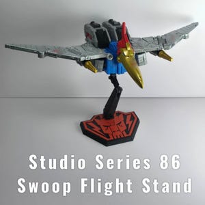 Op de afbeelding: Een speelgoedvliegtuig met grijze vleugels, een blauw en grijs lichaam en een gouden snavel wordt tentoongesteld op een zwarte standaard met een rode en zwarte basis. De tekst "Studio Series 86 Swoop Flight Stand" staat eronder.