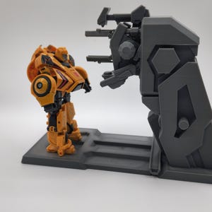 Puede incluir: Un juguete robot amarillo y negro se encuentra frente a un juego de pl&aacute;stico gris con una torreta de ca&ntilde;&oacute;n. El robot est&aacute; en posici&oacute;n con los brazos a los lados. El juego tiene una plataforma elevada y una torreta de ca&ntilde;&oacute;n con tres ca&ntilde;ones.