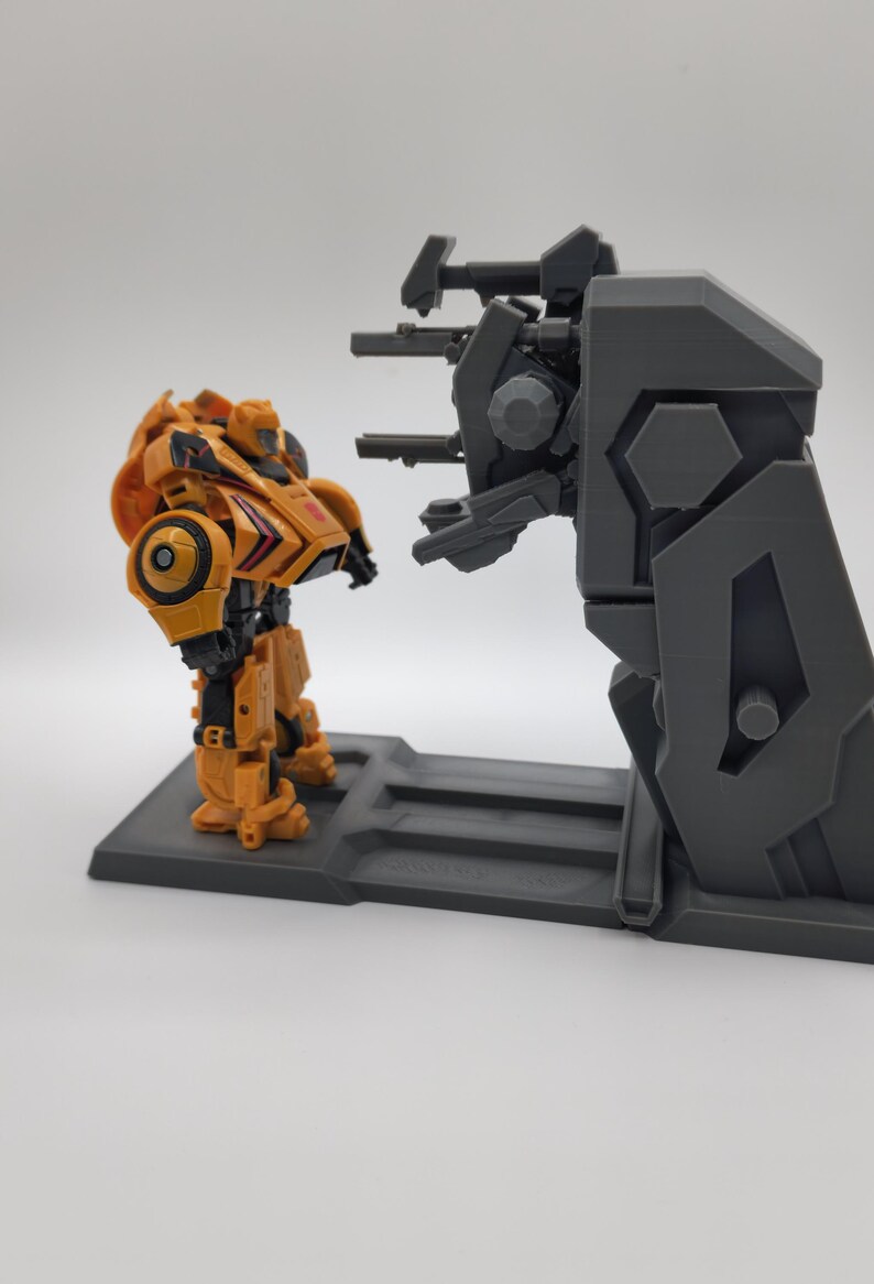 Puede incluir: Un juguete robot amarillo y negro se encuentra frente a un juego de pl&aacute;stico gris con una torreta de ca&ntilde;&oacute;n. El robot est&aacute; en posici&oacute;n con los brazos a los lados. El juego tiene una plataforma elevada y una torreta de ca&ntilde;&oacute;n con tres ca&ntilde;ones.