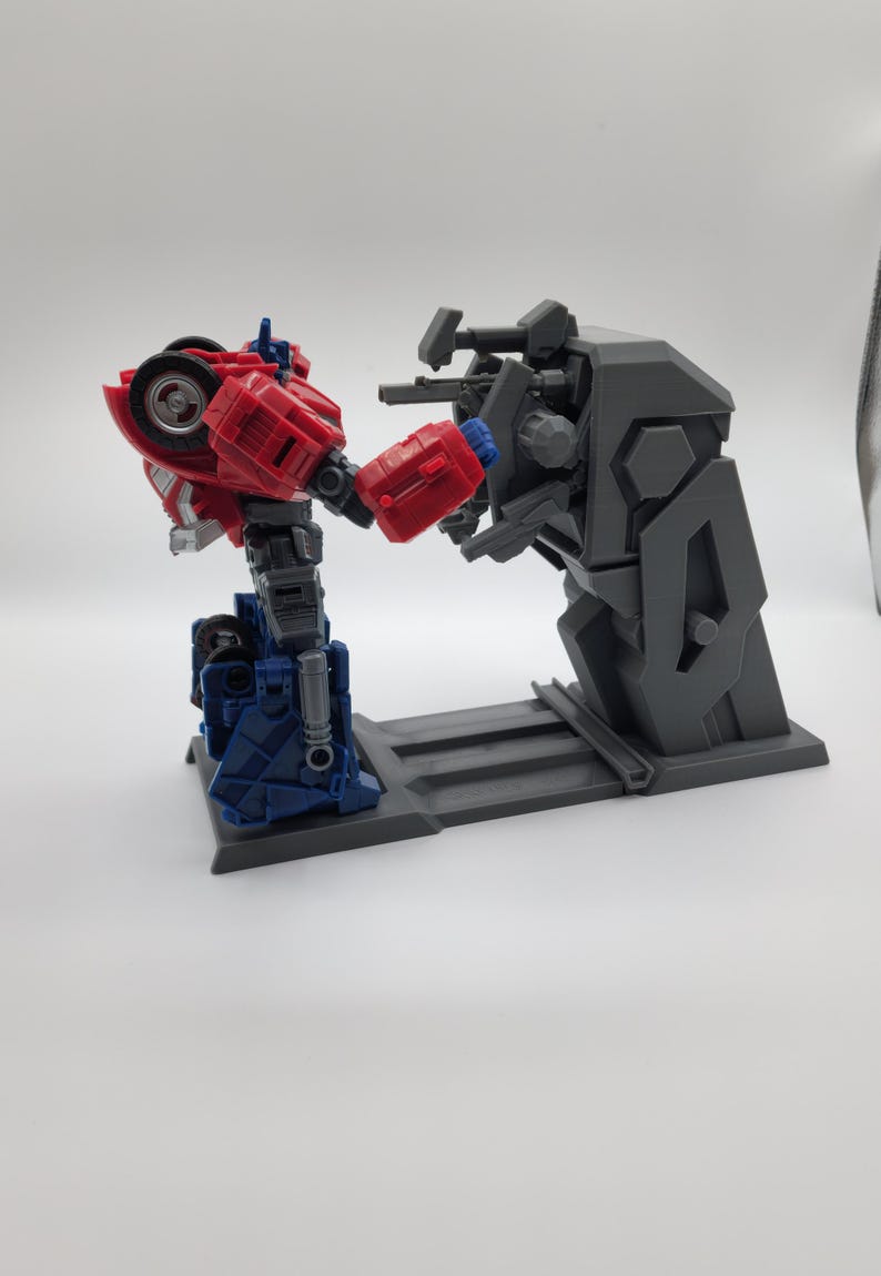 Puede incluir: Una figura de acci&oacute;n de robot rojo y azul est&aacute; en posici&oacute;n de combate, frente a un soporte de robot de pl&aacute;stico gris con m&uacute;ltiples armas. El soporte tiene una base gris con una superficie texturizada.