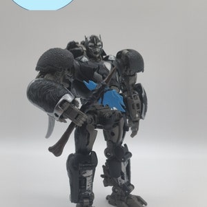 May include: A black and silver robot toy with a blue axe. The robot has a gorilla-like head and is holding the axe in its right hand. The toy is labelled "SS106 Primal X Kong Axe".