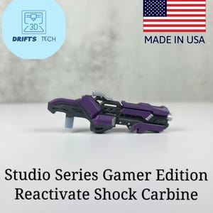 Può includere: Un fucile Reactivate Shock Carbine Gamer Edition della serie Studio, viola e grigio. L'immagine include il testo "Made in USA" e un logo Drifts Tech.