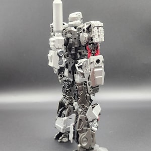 Peut inclure: Figurine d'action grise et noire de Megatron de la série Transformers Studio. La figurine a un canon d'épaule et est étiquetée SS109. L'image comprend également le texte "Transformers Studio Series" et "Drifts Tech".