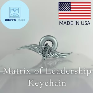 May include: A silver-colored keychain with a unique design, featuring a ring and stylized wings. The text "Matrix of Leadership Keychain" is displayed below the product. The image also includes the text "Made in USA" and the American flag.