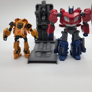 Puede incluir: Un robot amarillo y negro se encuentra junto a una plataforma gris con un robot rojo y azul de pie en la plataforma. El robot rojo y azul tiene un pecho gris y una cabeza negra.