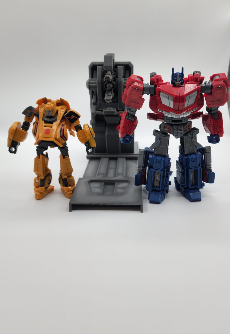 Puede incluir: Un robot amarillo y negro se encuentra junto a una plataforma gris con un robot rojo y azul de pie en la plataforma. El robot rojo y azul tiene un pecho gris y una cabeza negra.