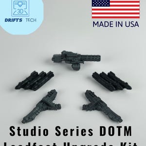 May include: A collection of dark gray Studio Series DOTM Leadfoot Upgrade Kit parts. The image includes a large gun and several smaller weapon attachments. The American flag and "MADE IN USA" text are visible in the upper right corner.
