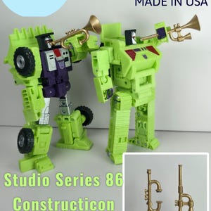 Puede incluir: Dos figuras de robots verdes y morados con trompetas doradas. La imagen incluye el texto "Studio Series 86 Constructicon Upgrade Kit Trumpets" y el texto "Made in USA".
